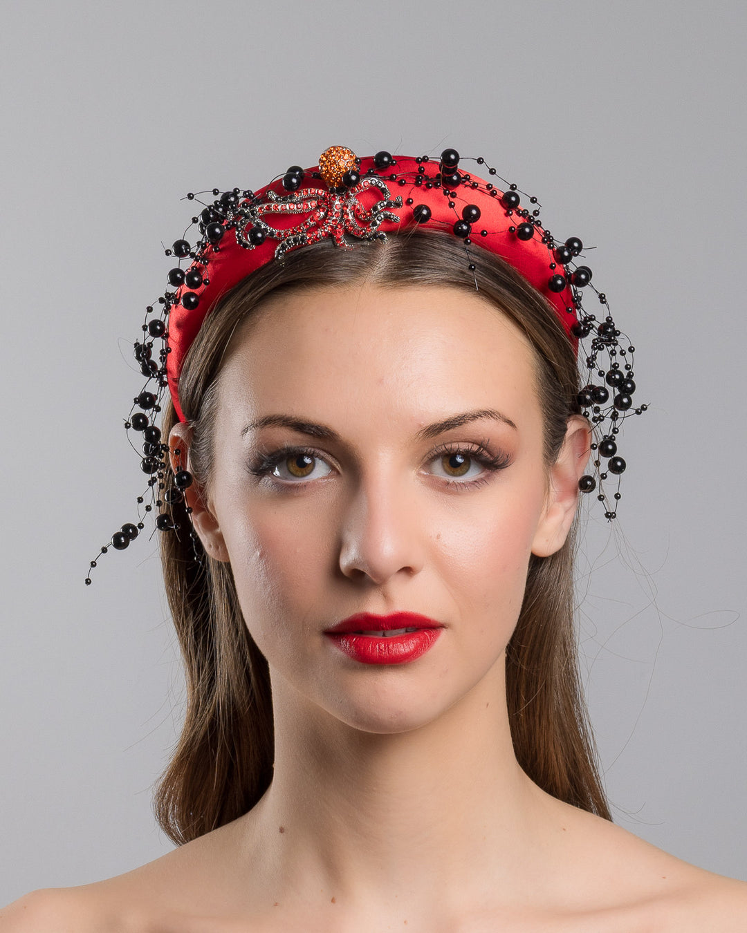 Model showcasing the front side close up of bright red silk sateen headband, decorated with crystal embellish octopus brooch and multi size black loose beads. Made by Merve Bayindir Millinery.