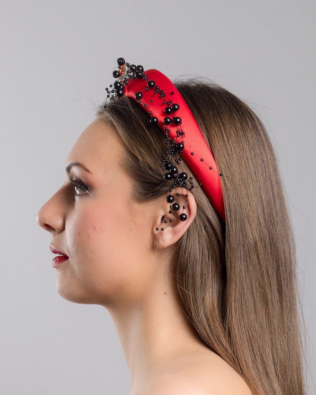 Model showcasing the left side close up of bright red silk sateen headband, decorated with crystal embellish octopus brooch and multi size black loose beads. Made by Merve Bayindir Millinery.