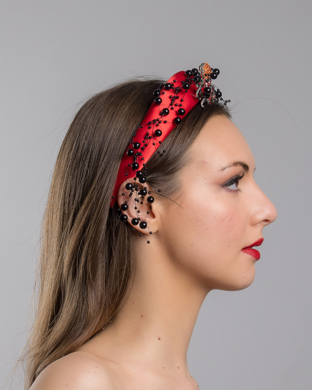 Model showcasing the right side close up of bright red silk sateen headband, decorated with crystal embellish octopus brooch and multi size black loose beads. Made by Merve Bayindir Millinery.