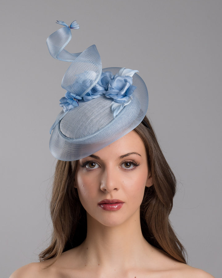 The model is showcasing the front side close up of the light blue button shaped fascinator. This unique fascinator is circled with light blue crinoline and is decorated with blue roses, shaded blue leaves and blue organza butterflies. Designed and made by Merve Bayindir Millinery.
