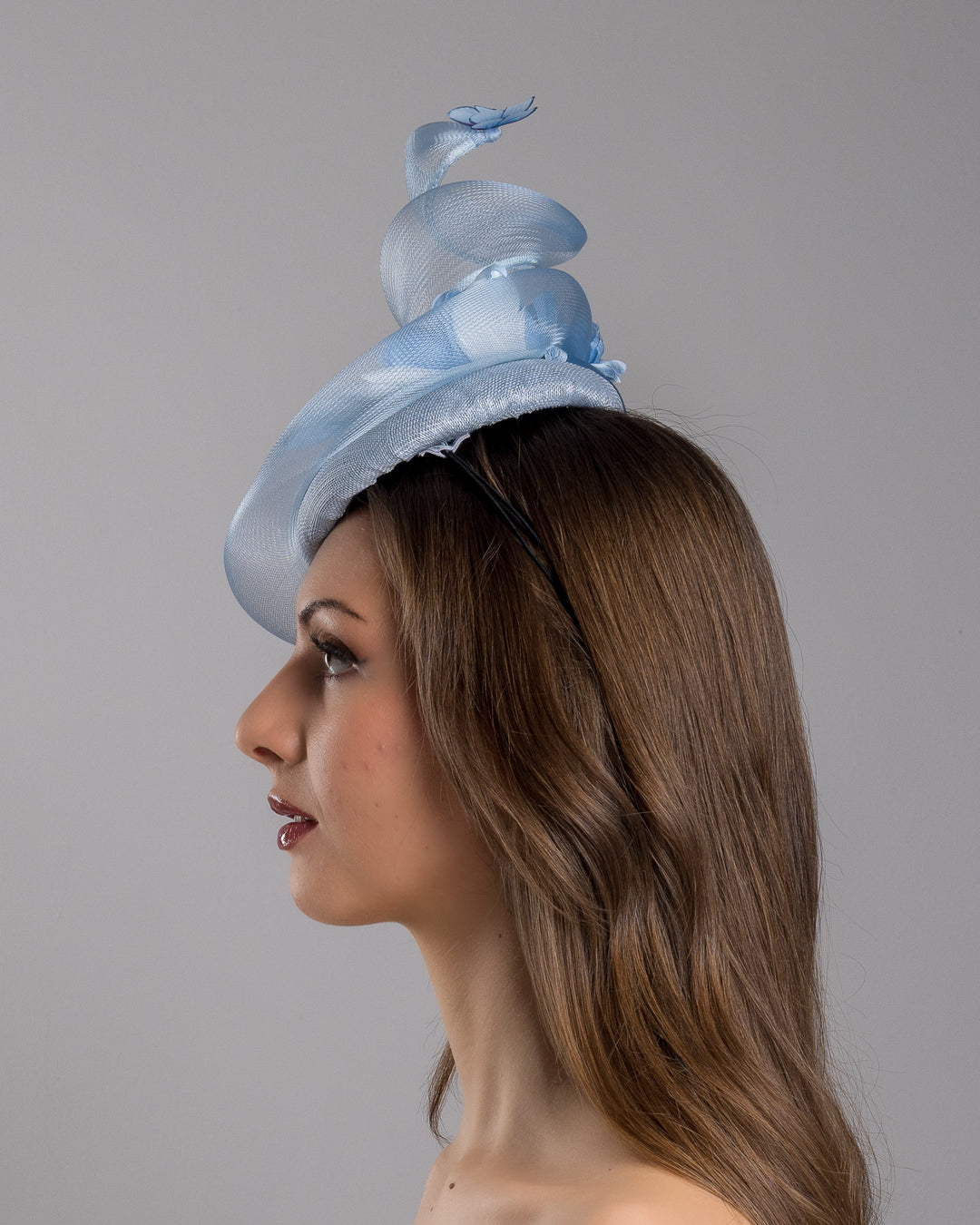 The model is showcasing the left side close up of the light blue button shaped fascinator. This unique fascinator is circled with light blue crinoline and is decorated with blue roses, shaded blue leaves and blue organza butterflies. Designed and made by Merve Bayindir Millinery.
