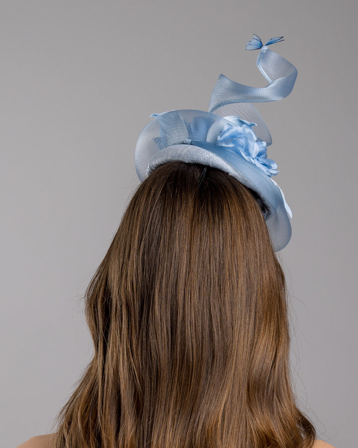 The model is showcasing the back side close up of the light blue button shaped fascinator. This unique fascinator is circled with light blue crinoline and is decorated with blue roses, shaded blue leaves and blue organza butterflies. Designed and made by Merve Bayindir Millinery.
