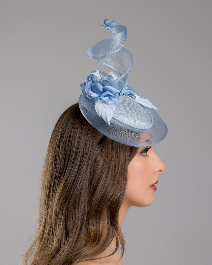 The model is showcasing the right side close up of the light blue button shaped fascinator. This unique fascinator is circled with light blue crinoline and is decorated with blue roses, shaded blue leaves and blue organza butterflies. Designed and made by Merve Bayindir Millinery.
