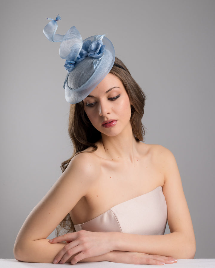 The  female model is posing, wearing Onyx,the light blue button shaped fascinator. This unique fascinator is circled with light blue crinoline and is decorated with blue roses, shaded blue leaves and blue organza butterflies. Designed and made by Merve Bayindir Millinery.
