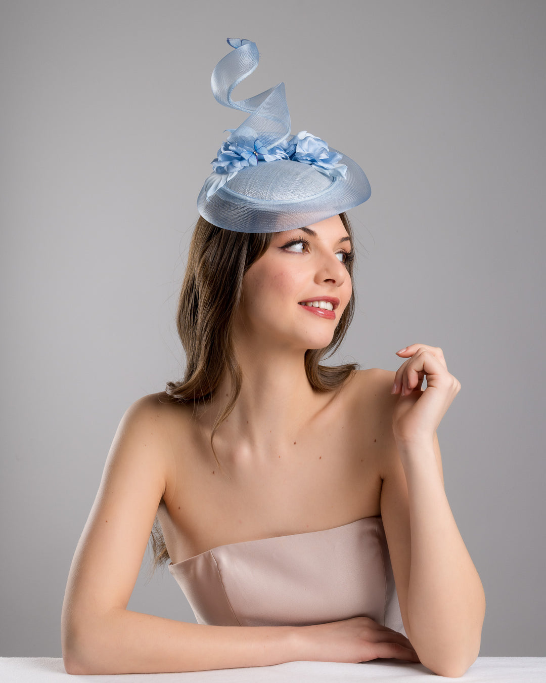 The  female model is posing, wearing Onyx,the light blue button shaped fascinator. This unique fascinator is circled with light blue crinoline and is decorated with blue roses, shaded blue leaves and blue organza butterflies. Designed and made by Merve Bayindir Millinery.

