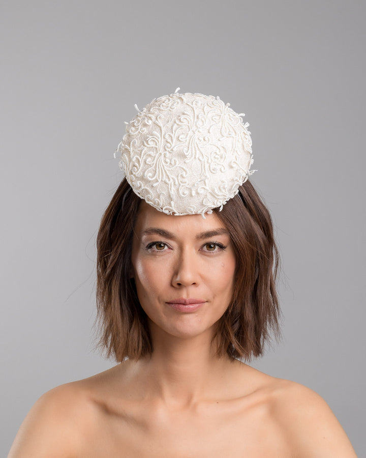 Model is showcasing the front side close up of double sided, white high top button style fascinator covered with sequins and lace with embroidered 3D swirls. Positioned on the top front centre of the head. Designed and made by Merve Bayindir Millinery.
