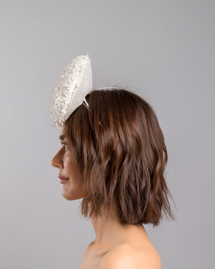 Model is showcasing the left side close up of double sided, white high top button style fascinator covered with sequins and lace with embroidered 3D swirls. Positioned on the top front centre of the head. Designed and made by Merve Bayindir Millinery.