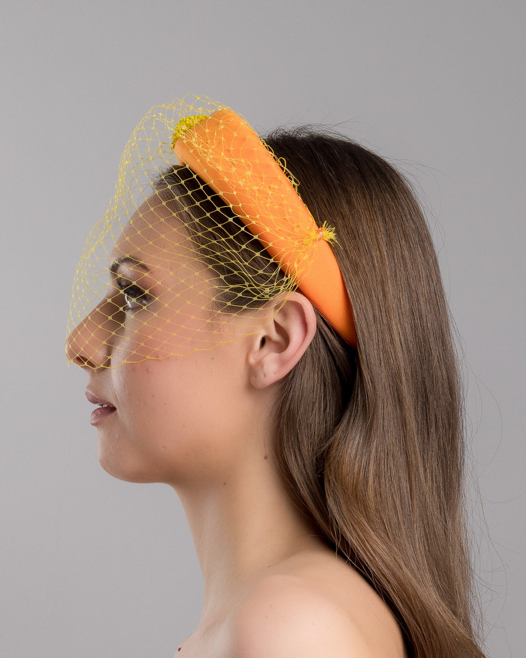 Model is showcasing the left side close up of high orange headband with yellow veil, and three yellow bead cluster. Made by Merve Bayindir Millinery.