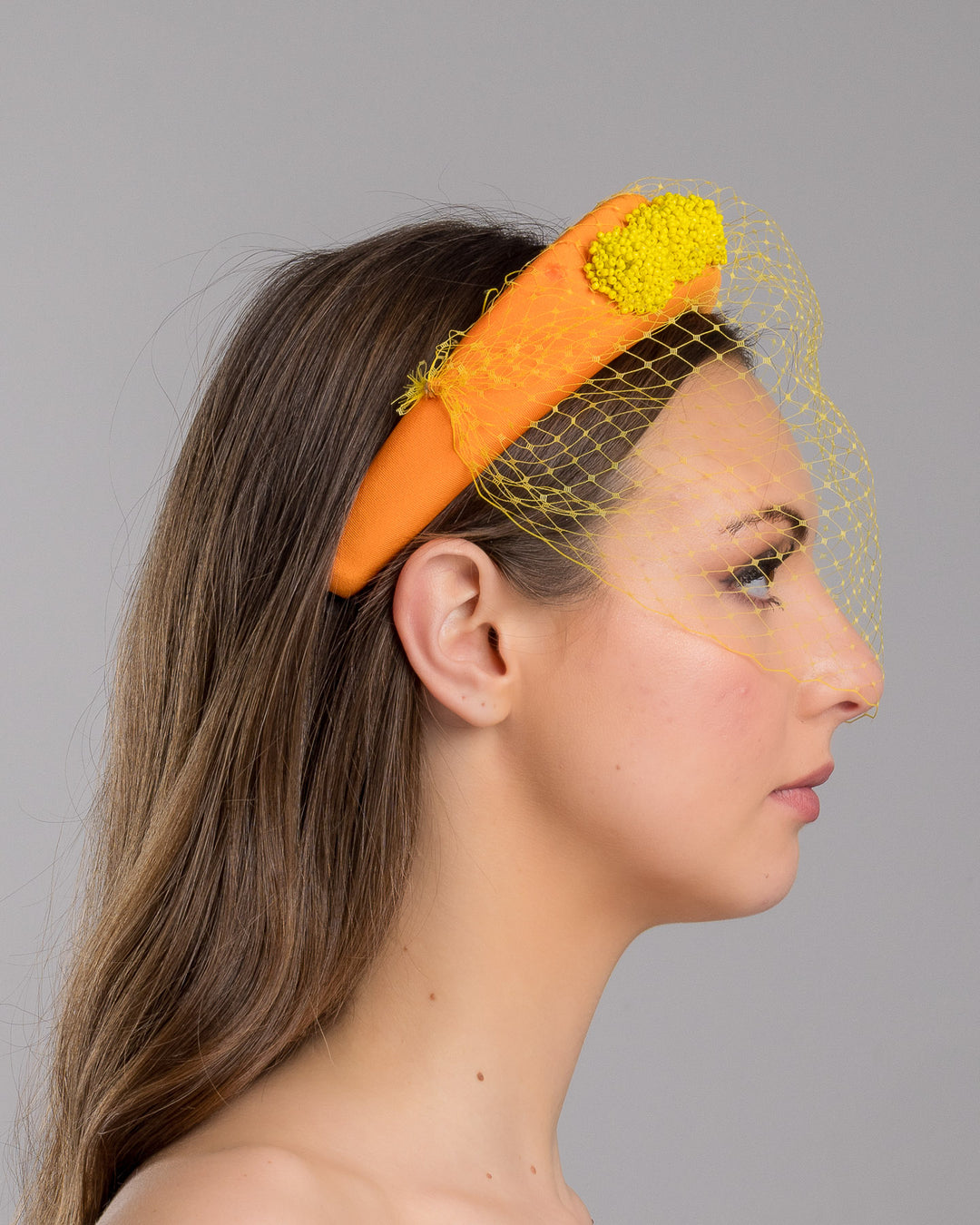 Model is showcasing the right side close up of high orange headband with yellow veil, and three yellow bead cluster. Made by Merve Bayindir Millinery.
