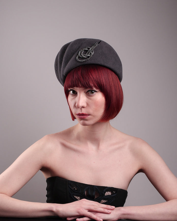 The model showcases the front left side of the Orion beret, made from deep grey wool felt, a classic vintage style shape is transformed by a sculptural knot in metallic gunmetal crystal rhinestone, resting near the temple like an insignia. Designed by Merve Bayindir.