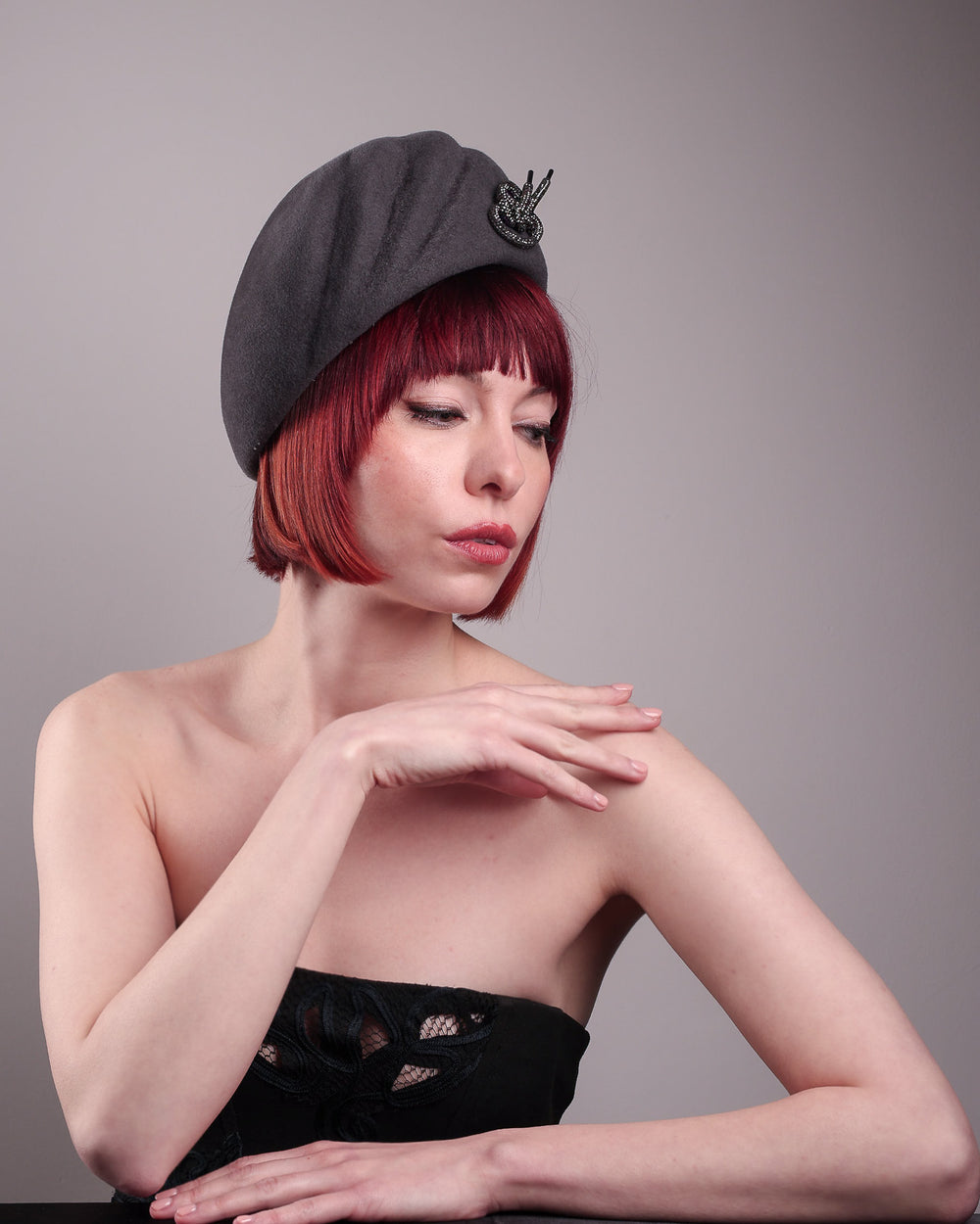 The model showcases the front of the Orion beret, made from deep grey wool felt, a classic vintage style shape is transformed by a sculptural knot in metallic gunmetal crystal rhinestone, resting near the temple like an insignia. Designed by Merve Bayindir.
