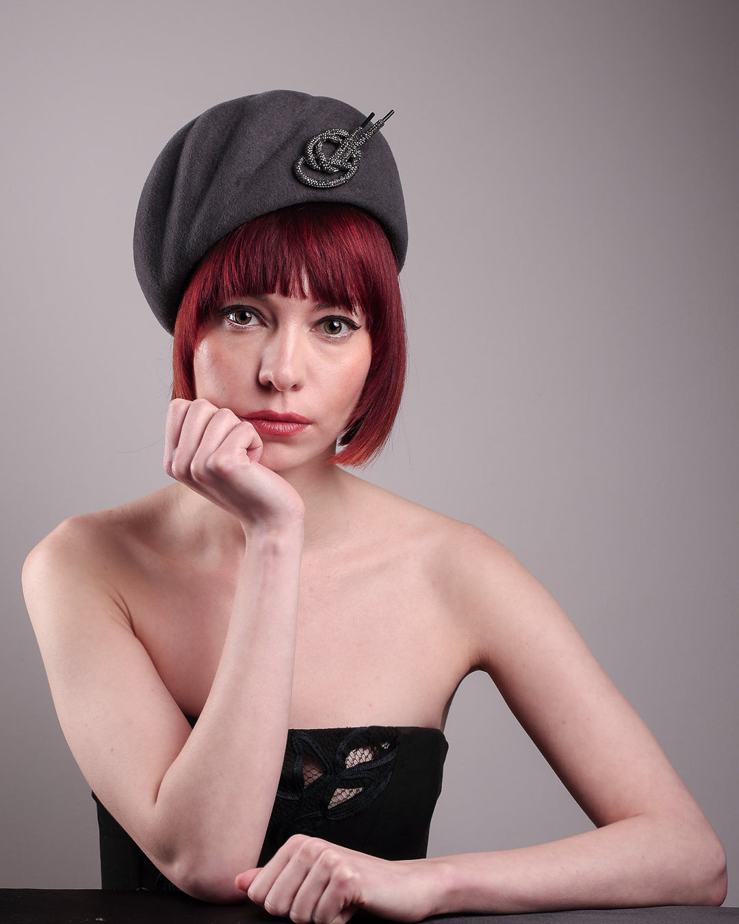 The model showcases the front of the Orion beret, made from deep grey wool felt, a classic vintage style shape is transformed by a sculptural knot in metallic gunmetal crystal rhinestone, resting near the temple like an insignia. Designed by Merve Bayindir.