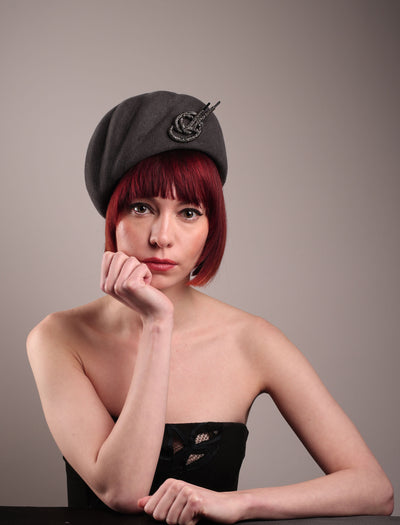 The model showcases the front of the Orion beret, made from deep grey wool felt, a classic vintage style shape is transformed by a sculptural knot in metallic gunmetal crystal rhinestone, resting near the temple like an insignia. Designed by Merve Bayindir.