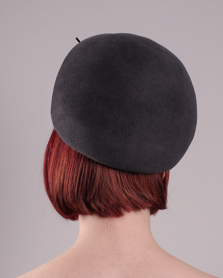 The model showcases the back of the Orion beret, made from deep grey wool felt, a classic vintage style shape is transformed by a sculptural knot in metallic gunmetal crystal rhinestone, resting near the temple like an insignia. Designed by Merve Bayindir.