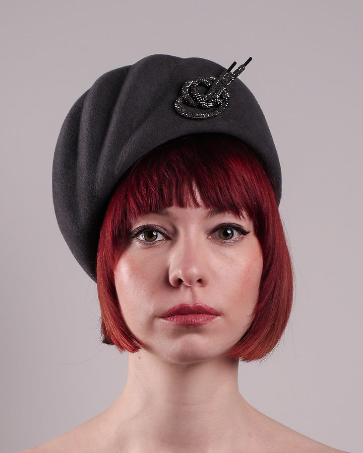 The model showcases the front close up of the Orion beret, made from deep grey wool felt, a classic vintage style shape is transformed by a sculptural knot in metallic gunmetal crystal rhinestone, resting near the temple like an insignia. Designed by Merve Bayindir.