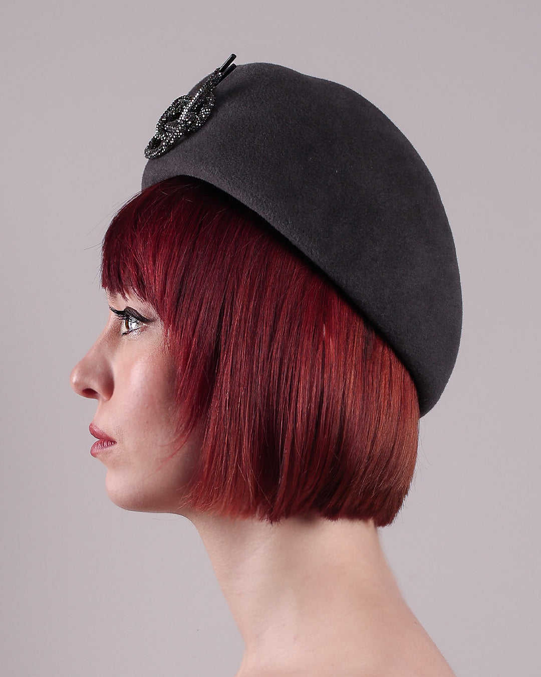 The model showcases the left close up of the Orion beret, made from deep grey wool felt, a classic vintage style shape is transformed by a sculptural knot in metallic gunmetal crystal rhinestone, resting near the temple like an insignia. Designed by Merve Bayindir.