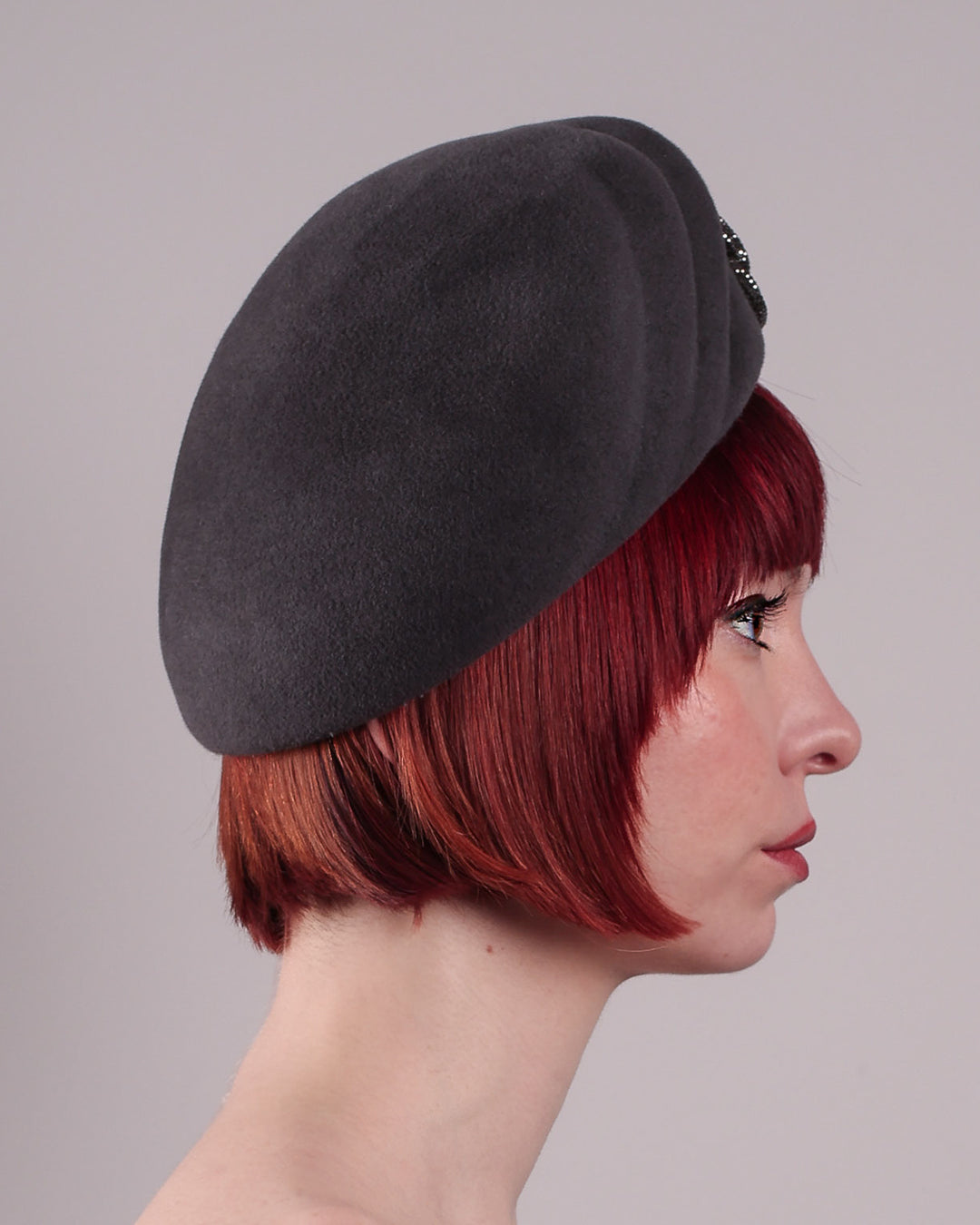 The model showcases the right side close up of the Orion beret, made from deep grey wool felt, a classic vintage style shape is transformed by a sculptural knot in metallic gunmetal crystal rhinestone, resting near the temple like an insignia. Designed by Merve Bayindir.