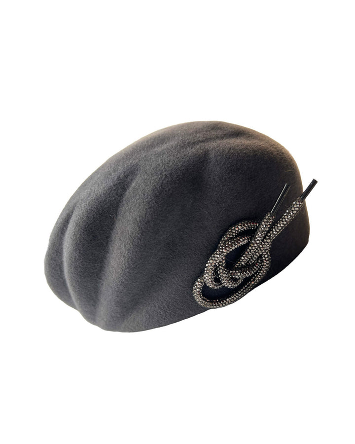 Still life picture of the Orion beret, made from deep grey wool felt, a classic vintage style shape is transformed by a sculptural knot in metallic gunmetal crystal rhinestone, resting near the temple like an insignia. Designed by Merve Bayindir.