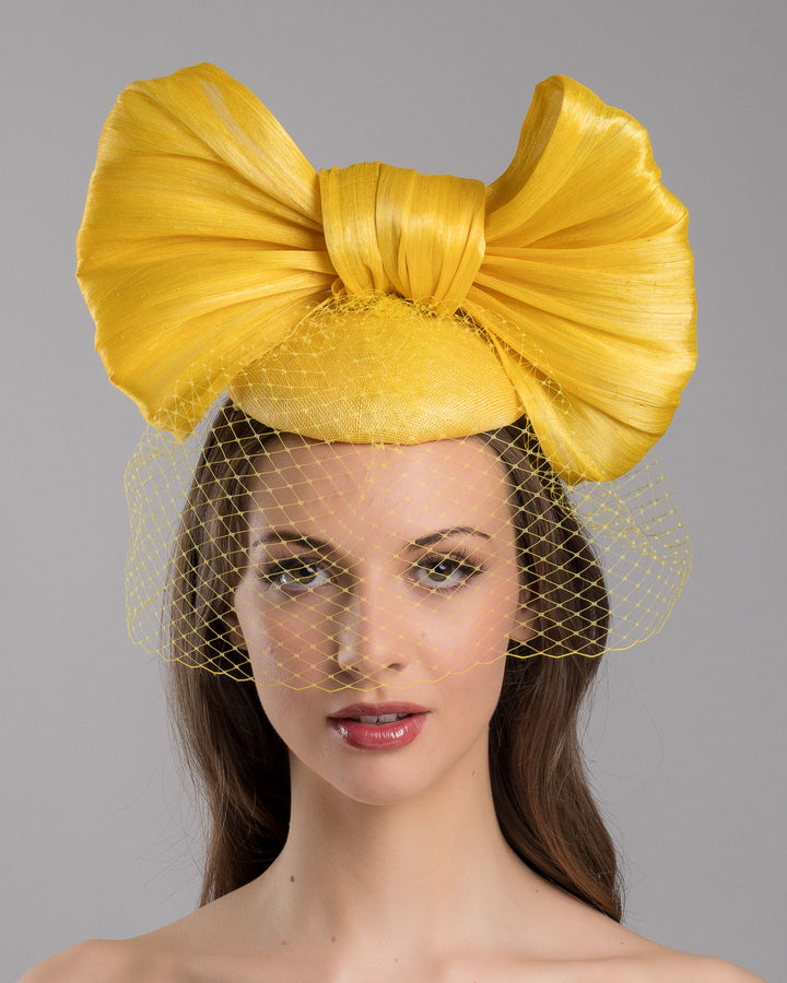 The model is showcasing the front side close up of the mustard yellow sinamay fascinator featuring an oversized structured bow, set on a matching base with a soft birdcage veil overlay and secured with a slim black headband. Designed and made by Merve Bayindir Millinery.