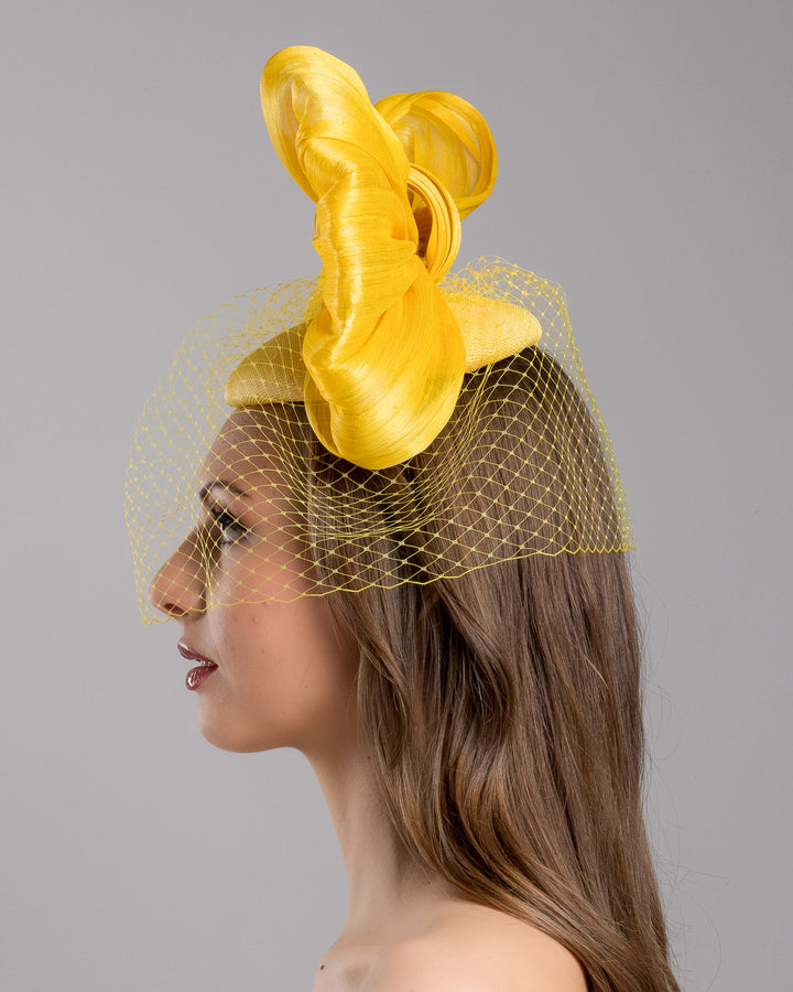 The model is showcasing the left side close up of the mustard yellow sinamay fascinator featuring an oversized structured bow, set on a matching base with a soft birdcage veil overlay and secured with a slim black headband. Designed and made by Merve Bayindir Millinery.