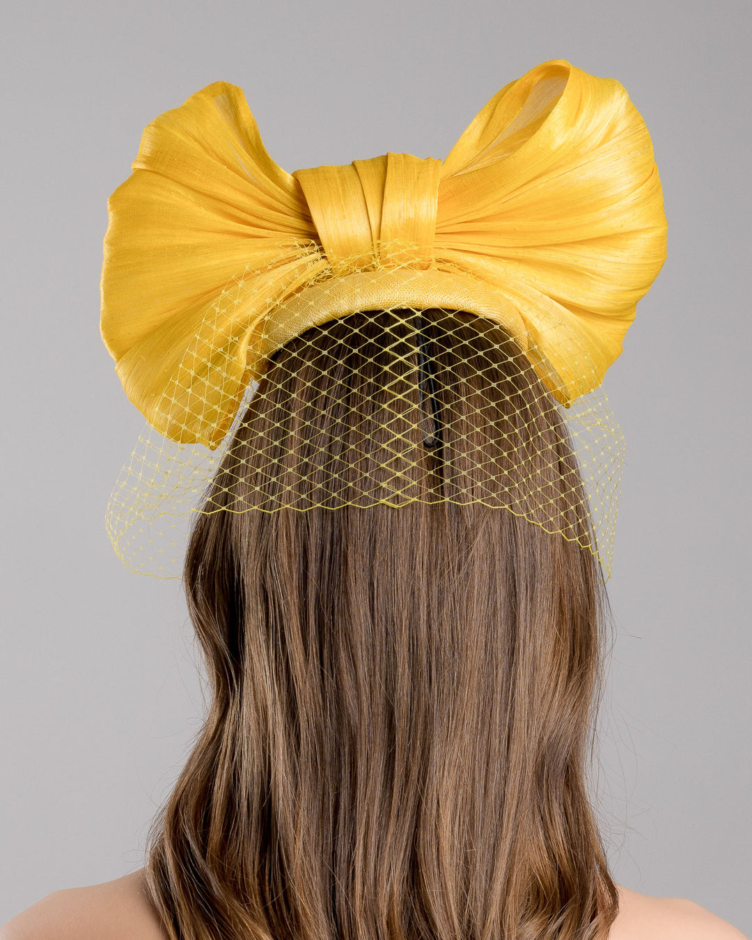 The model is showcasing the back side close up of the mustard yellow sinamay fascinator featuring an oversized structured bow, set on a matching base with a soft birdcage veil overlay and secured with a slim black headband. Designed and made by Merve Bayindir Millinery.