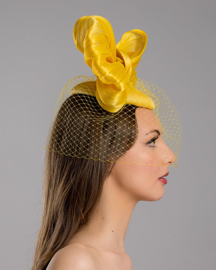 The model is showcasing the right side close up of the mustard yellow sinamay fascinator featuring an oversized structured bow, set on a matching base with a soft birdcage veil overlay and secured with a slim black headband. Designed and made by Merve Bayindir Millinery.
