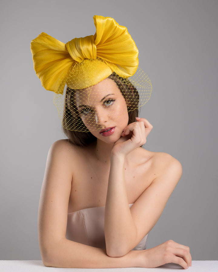 The female model is posing, wearing
Pat, the mustard yellow sinamay fascinator featuring an oversized structured bow, set on a matching base with a soft birdcage veil overlay and secured with a slim black headband. Designed and made by Merve Bayindir Millinery.