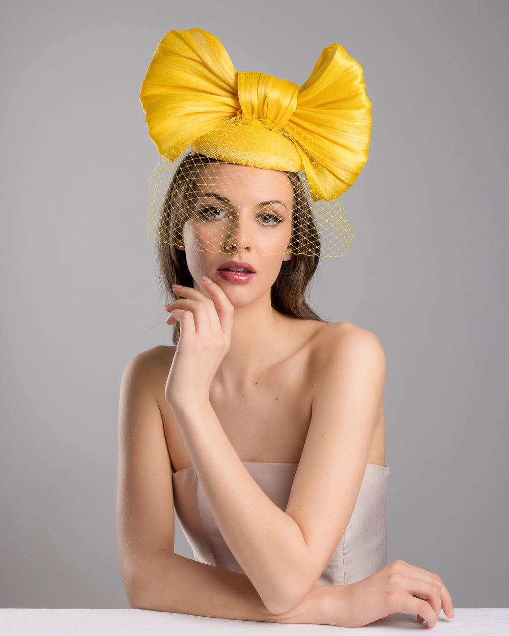 The female model is posing, wearing
Pat, the mustard yellow sinamay fascinator featuring an oversized structured bow, set on a matching base with a soft birdcage veil overlay and secured with a slim black headband. Designed and made by Merve Bayindir Millinery.