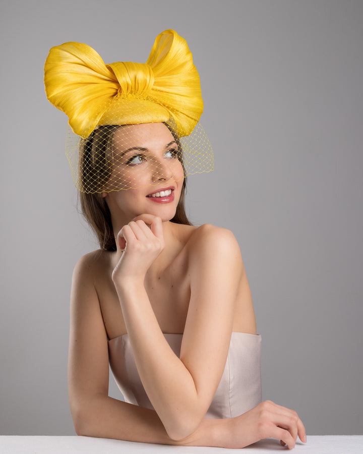 The female model is posing, wearing
Pat, the mustard yellow sinamay fascinator featuring an oversized structured bow, set on a matching base with a soft birdcage veil overlay and secured with a slim black headband. Designed and made by Merve Bayindir Millinery.