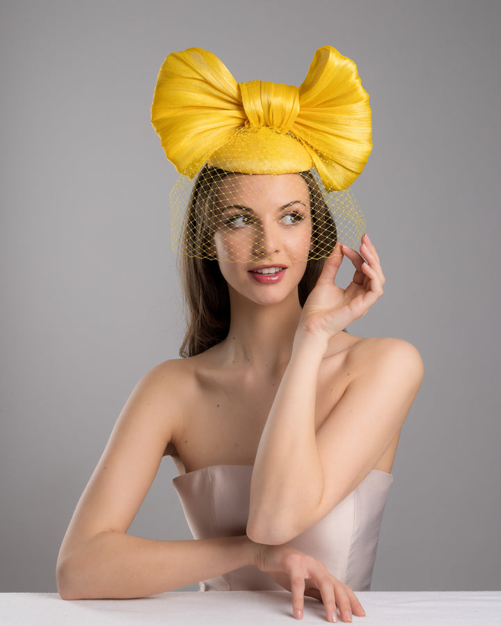 The female model is posing, wearing
Pat, the mustard yellow sinamay fascinator featuring an oversized structured bow, set on a matching base with a soft birdcage veil overlay and secured with a slim black headband. Designed and made by Merve Bayindir Millinery.