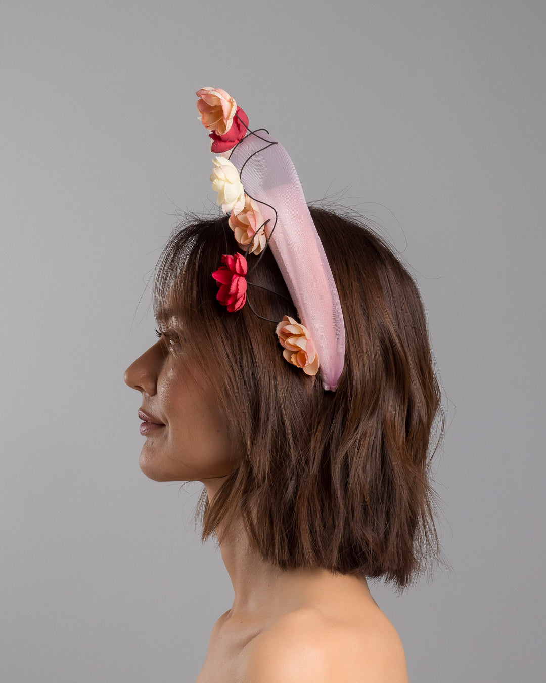 The model is showcasing the left side close up of the blush pink headband fascinator with a sheer pale halo frame, adorned with wired floral embellishments in ivory, peach, and red tones. Designed and Made by Merve Bayindir millinery.