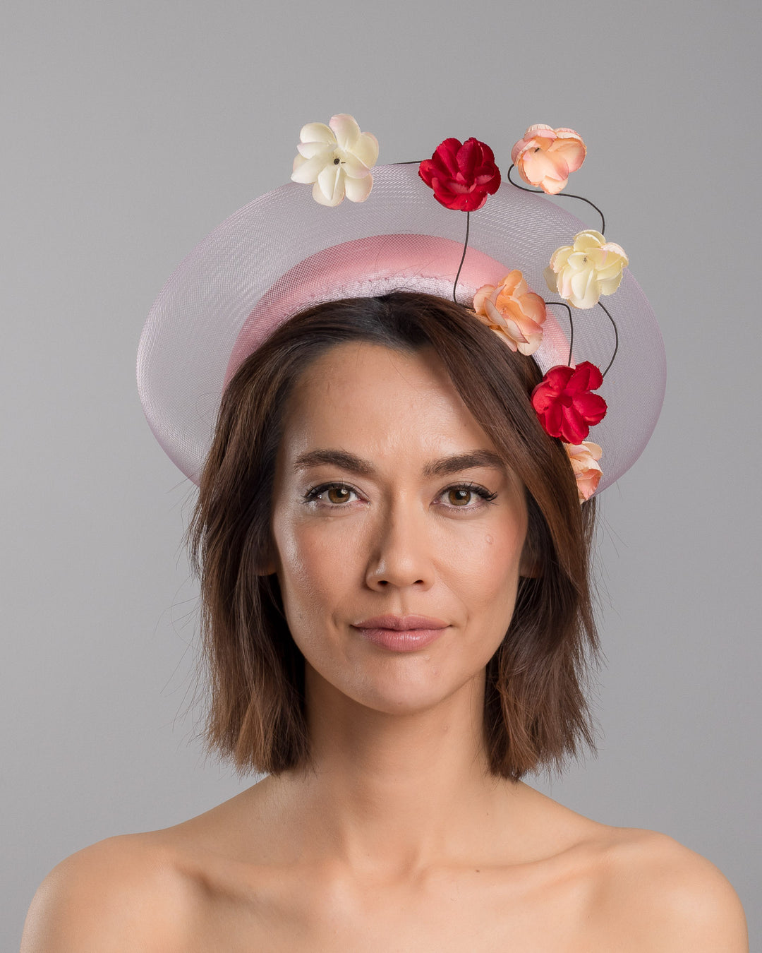 The model is showcasing the front side close up of the blush pink headband fascinator with a sheer pale halo frame, adorned with wired floral embellishments in ivory, peach, and red tones. Designed and Made by Merve Bayindir millinery.