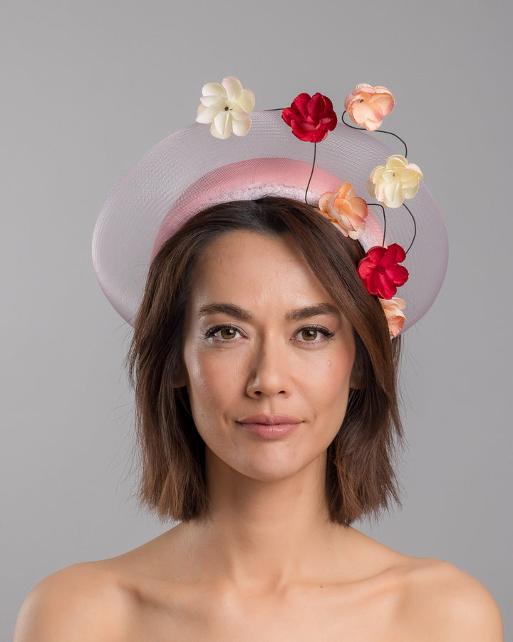 The model is showcasing the front side close up of the blush pink headband fascinator with a sheer pale halo frame, adorned with wired floral embellishments in ivory, peach, and red tones. Designed and Made by Merve Bayindir millinery.
