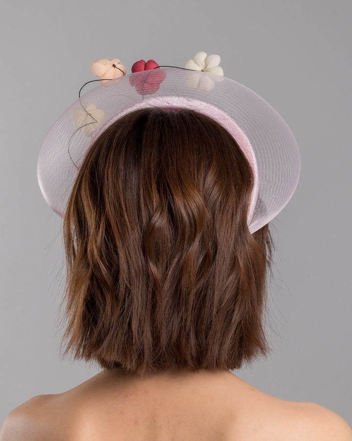 The model is showcasing the back side close up of the blush pink headband fascinator with a sheer pale halo frame, adorned with wired floral embellishments in ivory, peach, and red tones. Designed and Made by Merve Bayindir millinery.