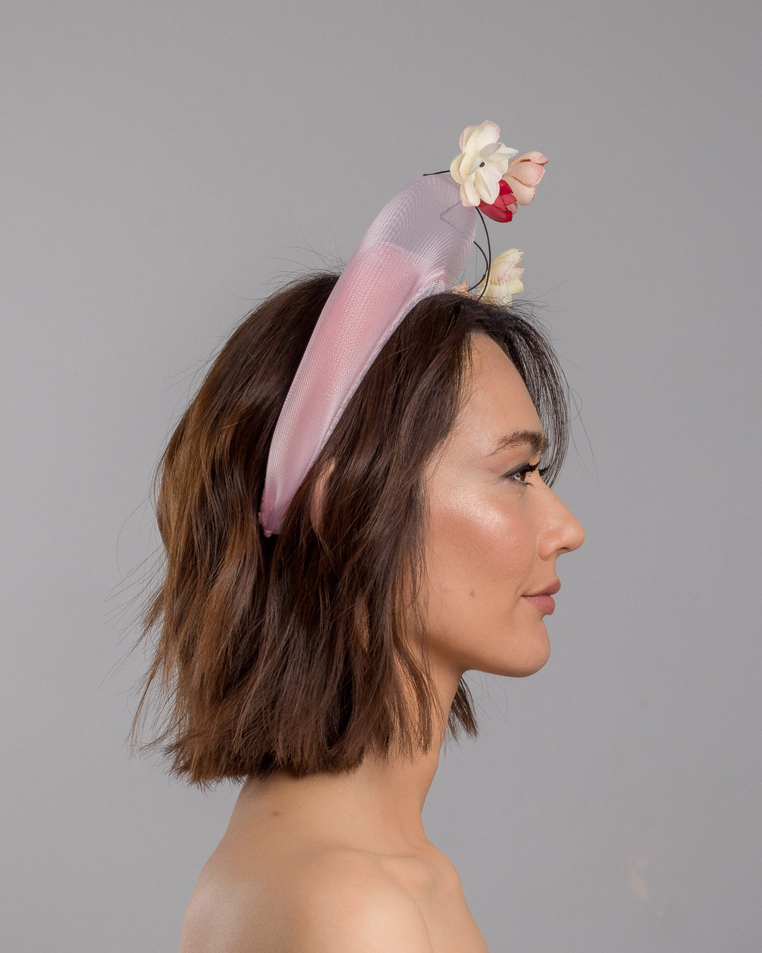 The model is showcasing the right side close up of the blush pink headband fascinator with a sheer pale halo frame, adorned with wired floral embellishments in ivory, peach, and red tones. Designed and Made by Merve Bayindir millinery.