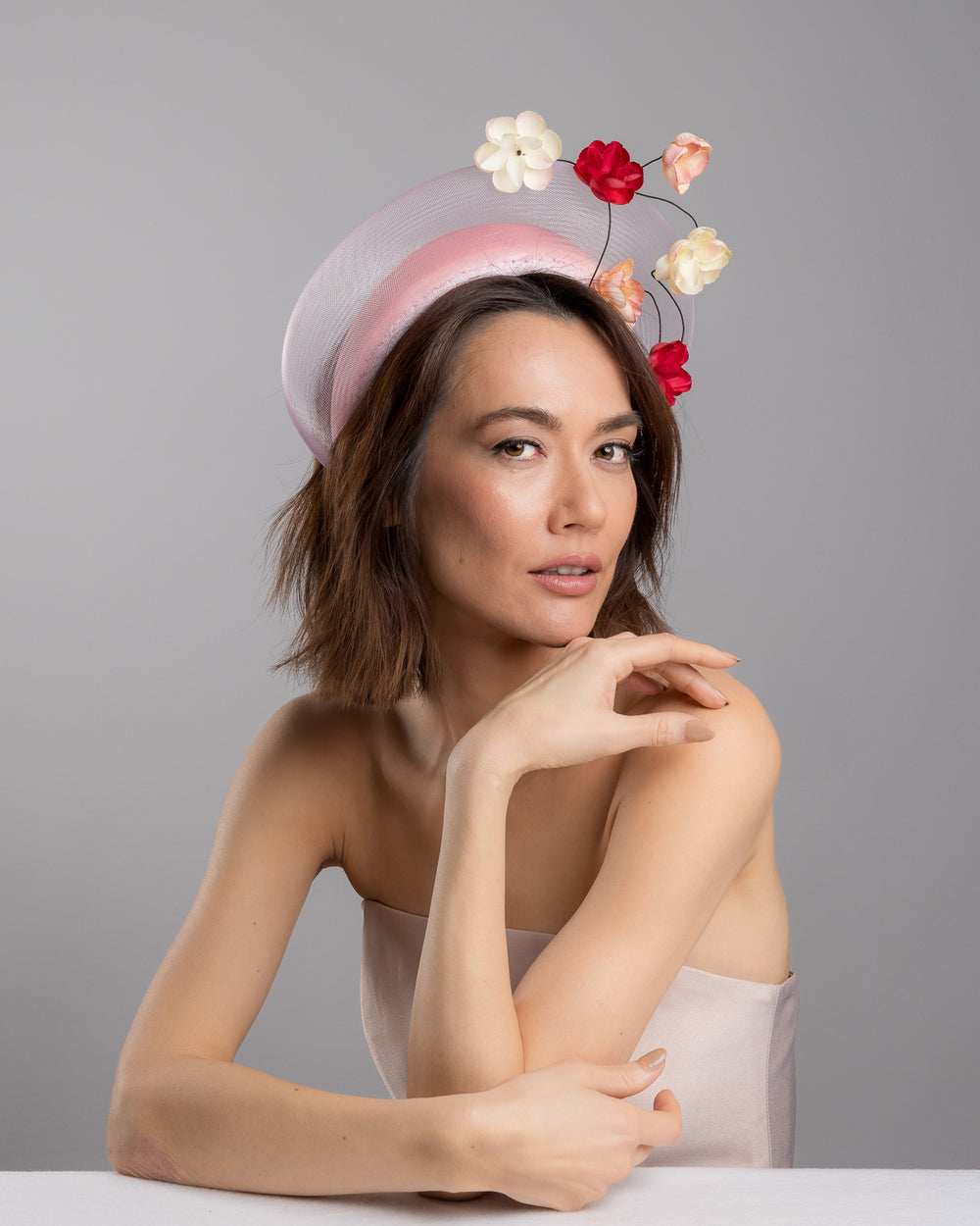 The female model is posing, wearing Petrae, the blush pink headband fascinator with a sheer pale halo frame, adorned with wired floral embellishments in ivory, peach, and red tones. Designed and Made by Merve Bayindir millinery.