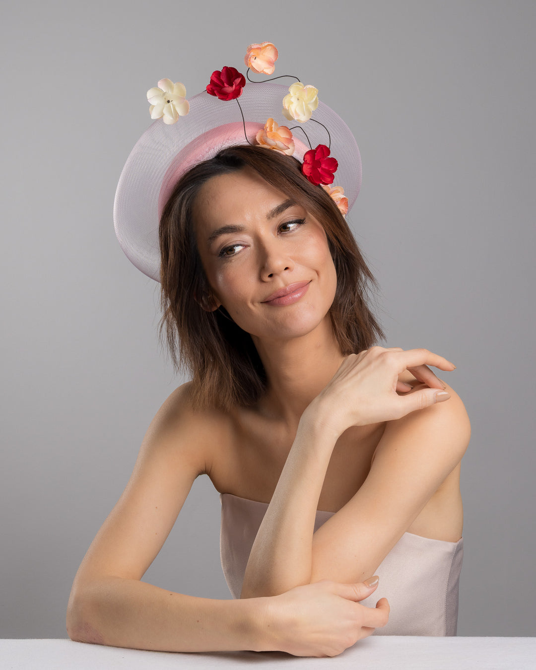 The female model is posing, wearing Petrae, the blush pink headband fascinator with a sheer pale halo frame, adorned with wired floral embellishments in ivory, peach, and red tones. Designed and Made by Merve Bayindir millinery.