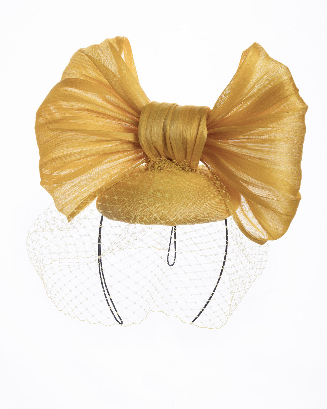 Mustard yellow sinamay fascinator featuring an oversized structured bow, set on a matching base with a soft birdcage veil overlay and secured with a slim black headband. Designed and made by Merve Bayindir Millinery.

