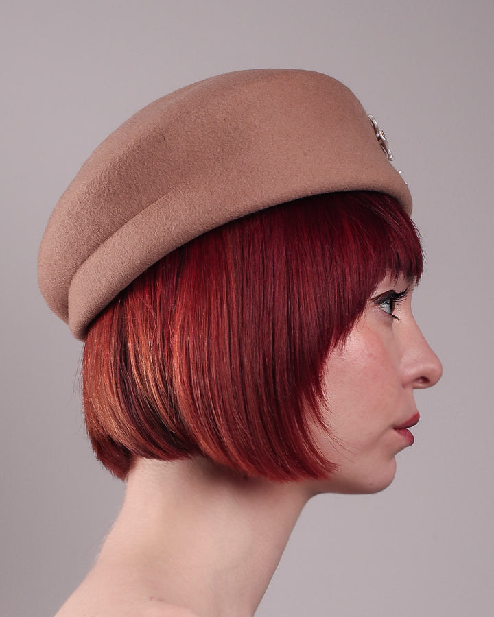 The female model is wearing and showcasing the right side close up of a camel-coloured felt beret with matte white-coloured metal flowers. The beret has a layer on the right side which the wearer also tilts towards.. Designed by Merve Bayindir.