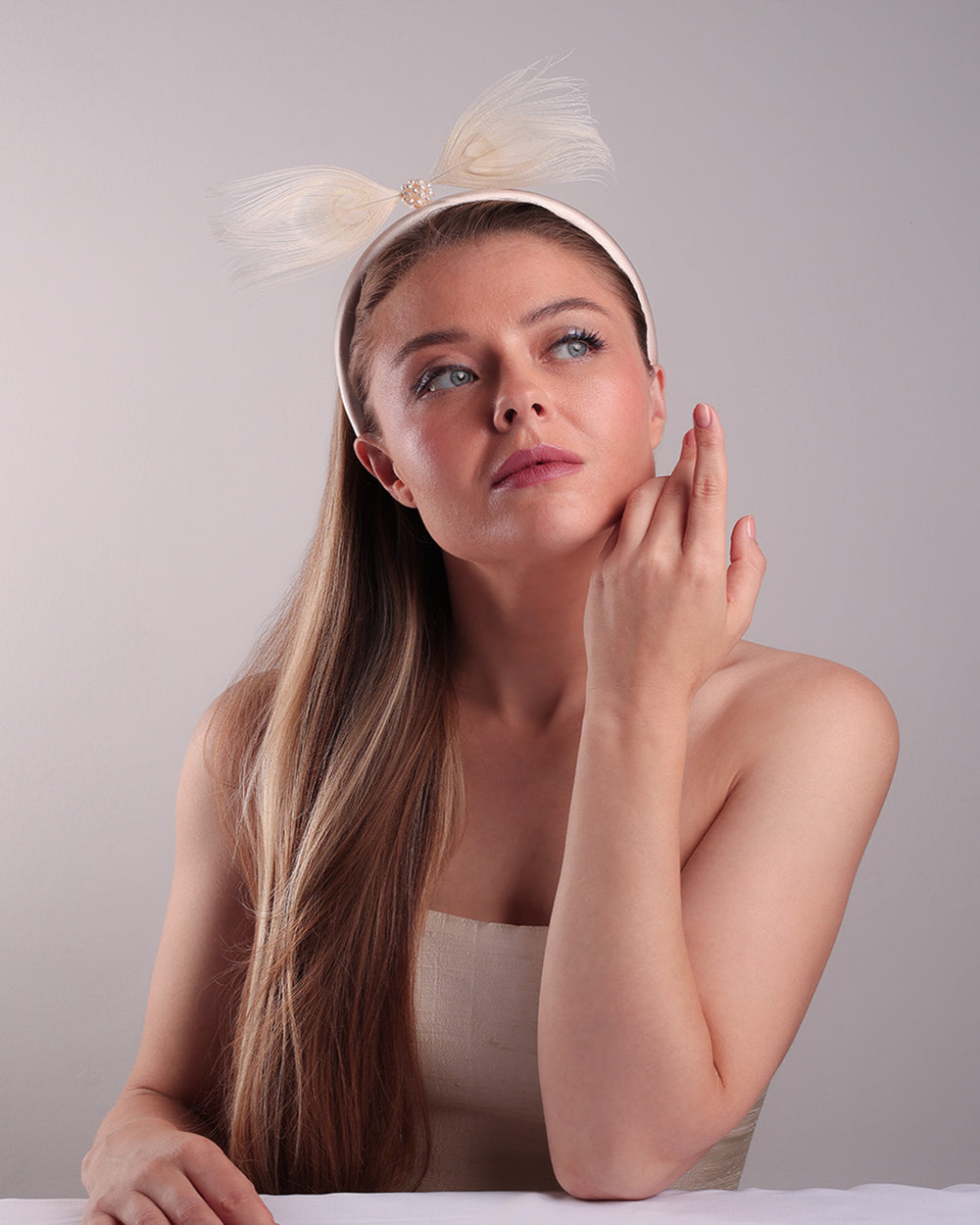 The model is showcasing the front of a pinkish ivory beige semi-high silk headband decorated with two ivory peacock feathers placed like bow centred with pearl cluter.