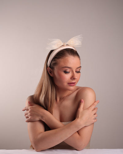 The model is showcasing the top front of a pinkish ivory beige semi-high silk headband decorated with two ivory peacock feathers placed like bow centred with pearl cluter.