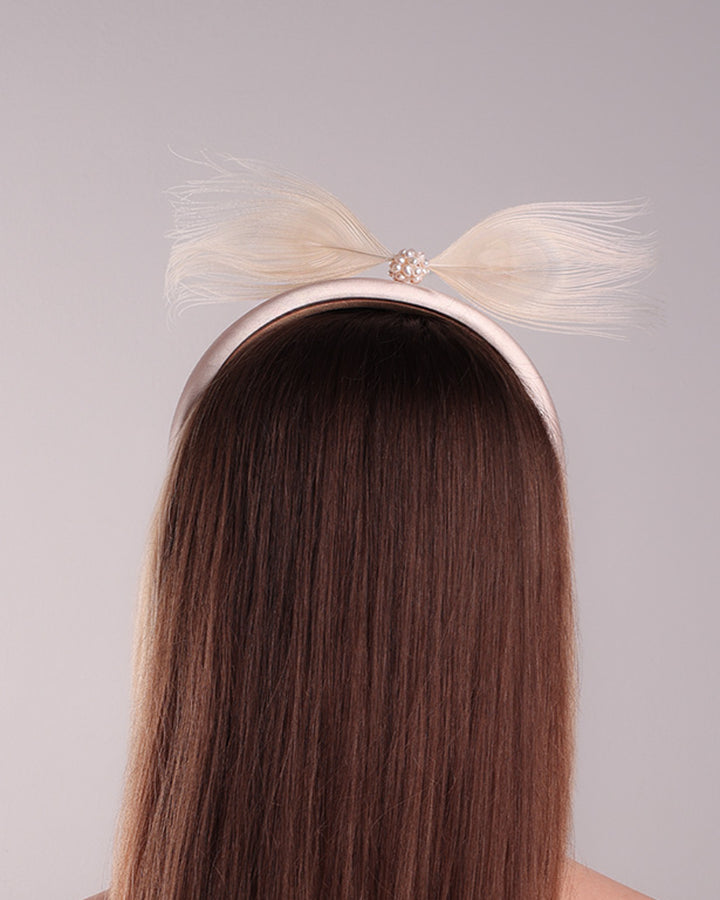 The model is showcasing the back of a pinkish ivory beige semi-high silk headband decorated with two ivory peacock feathers placed like bow centred with pearl cluter.