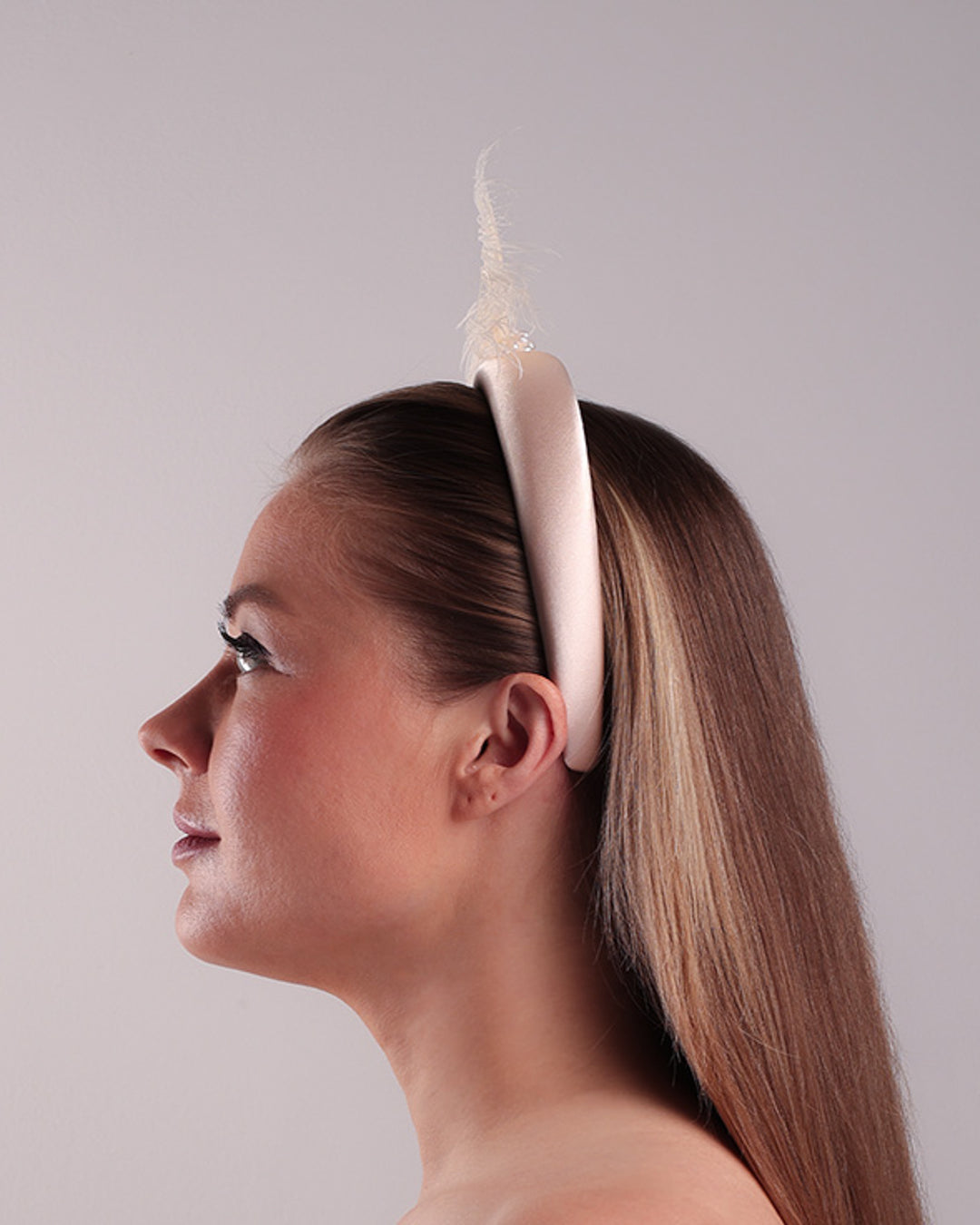 The model is showcasing the left side of a pinkish ivory beige semi-high silk headband decorated with two ivory peacock feathers placed like bow centred with pearl cluter.
