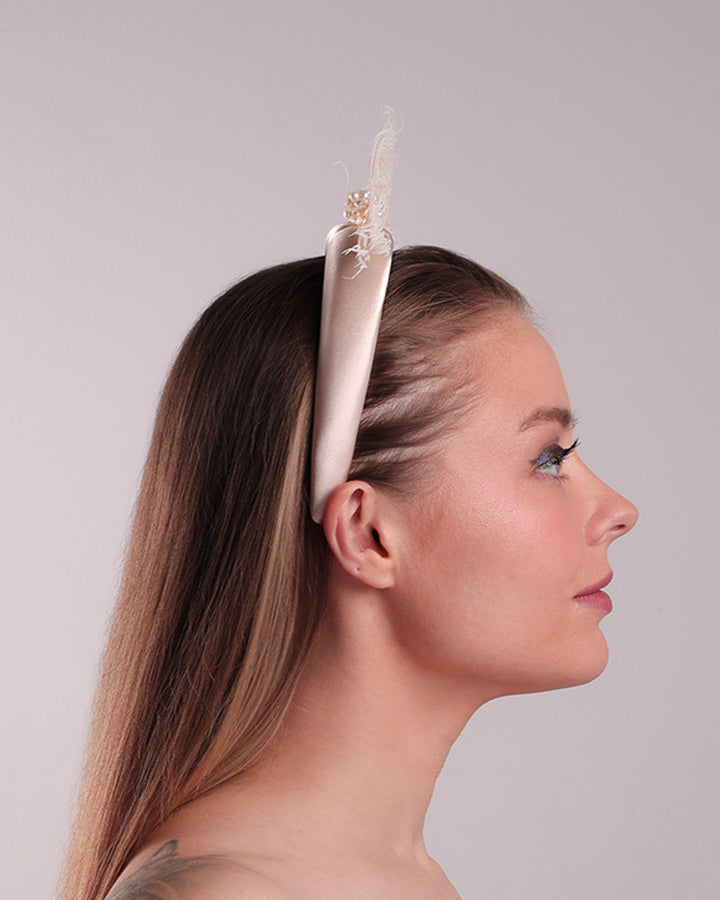 The model is showcasing the right side of a pinkish ivory beige semi-high silk headband decorated with two ivory peacock feathers placed like bow centred with pearl cluter.