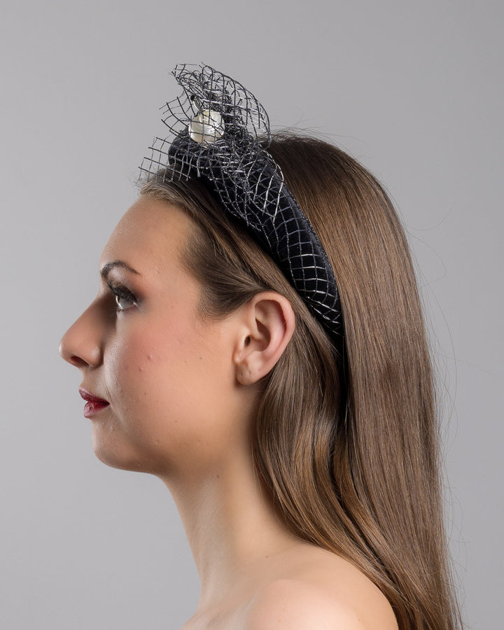 Model is showcasing the left side close up of Black Velvet High Headband, covered with a silver black mix net fabric. It is embellished with a net fabric flower and a mother of pearl detailed snail brooch. Made by Merve Bayindir Millinery.