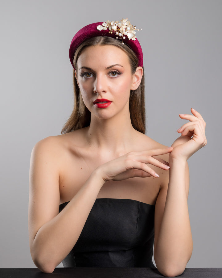 Model is posing wearing the Permissio a high burgundy velvet headband decorated with pearl coloured wired bead and resin flowers. Made by Merve Bayindir millinery.
