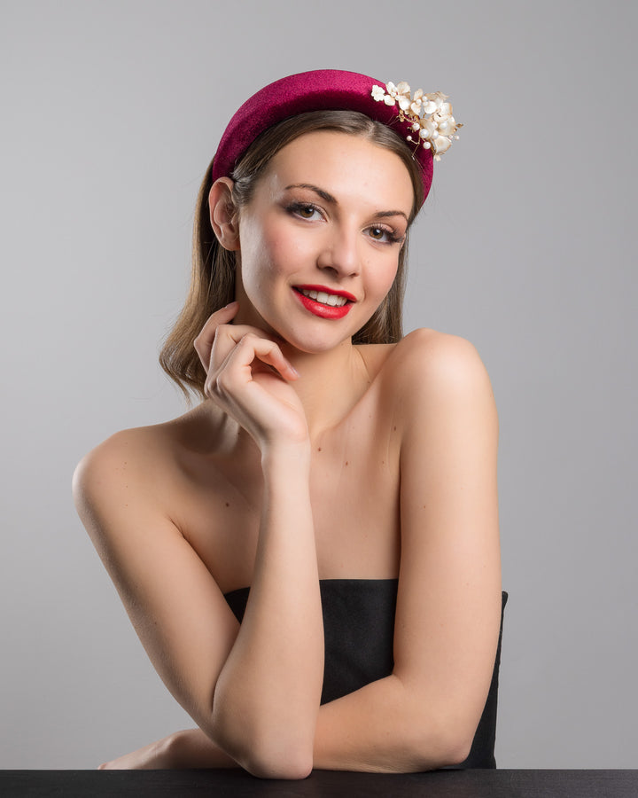 Model is posing wearing the Permissio a high burgundy velvet headband decorated with pearl coloured wired bead and resin flowers. Made by Merve Bayindir millinery.