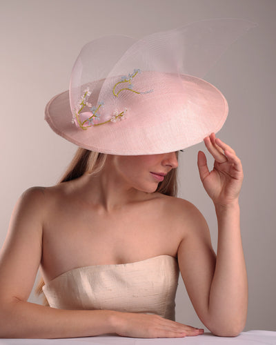 Model is showcasing the right detail of a wide brim hat named Petal Canopy. A large size pink oval pink wide-brim hat decorated with a crinoline ribbon tide on the right side of the hat is looking like flying towards the left. Right edge of the crinoline, and part of the front piece of the crinoline is decorated with flowers shaped beads.