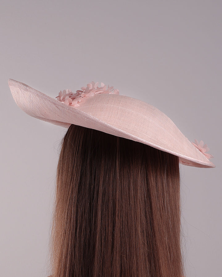 Model is showcasing the back of the medium size pink saucer hat, curved towards the left side and tilted to the right side, is decorated with multi petal pink silk sateen flowers, from top left side to right side. Centre of the flowers are embellished with pink pearls.