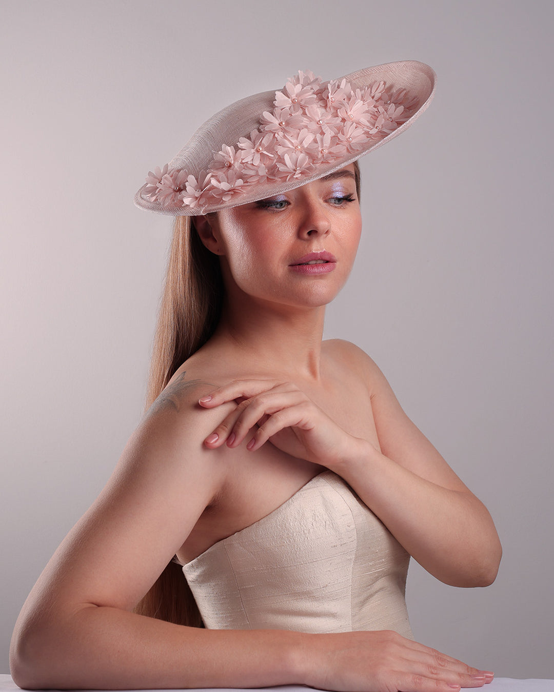 Model is showcasing the front right side of the medium size pink saucer hat, curved towards the left side and tilted to the right side, is decorated with multi petal pink silk sateen flowers, from top left side to right side. Centre of the flowers are embellished with pink pearls.